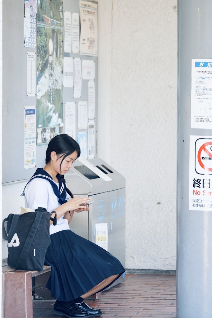 A young Japanese girl in school uniform sits indoors using a smartphone.
